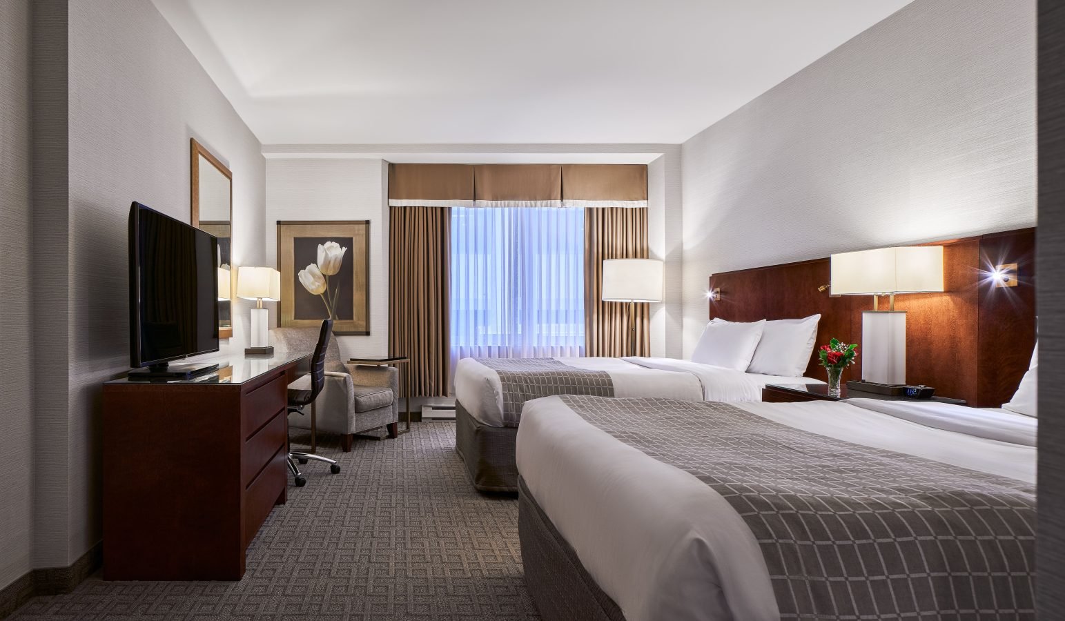 A Lord Elgin Hotel room featuring two double beds, a grey chair, a wooden dresser with a flat screen TV, and floor to ceiling windows.