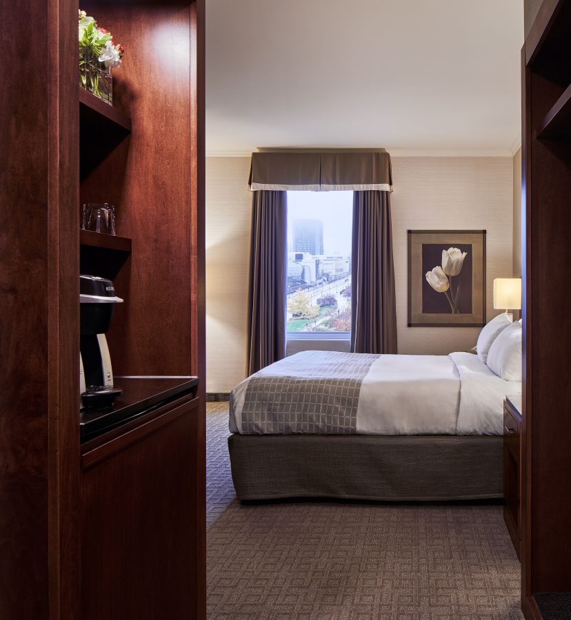A Lord Elgin Hotel room featuring a King-sized bed, a closet with wooden furnishings and a coffee maker. There is floor to ceiling windows and a framed picture of a white flower on the wall in the background.