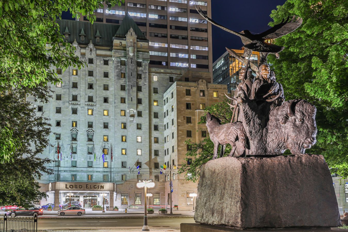 An outdoor view of the Lord Elgin Hotel at nighttime, from behind the statue in Confederation Park, just across the street. There is a large eagle, two people, and a wolf depicted in the statue.