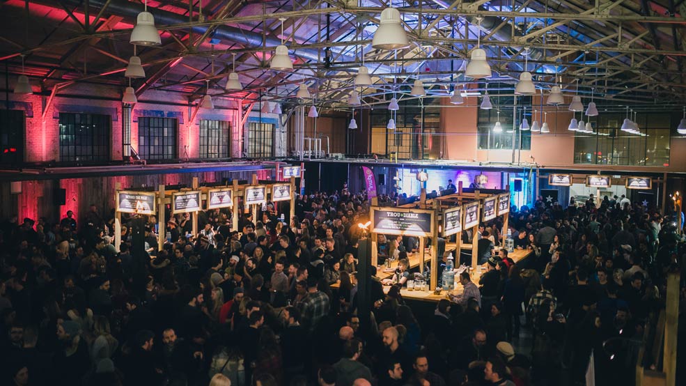 A crowd of people indoors at a beer tasting festival, with various stands made out of wood serving beverages.