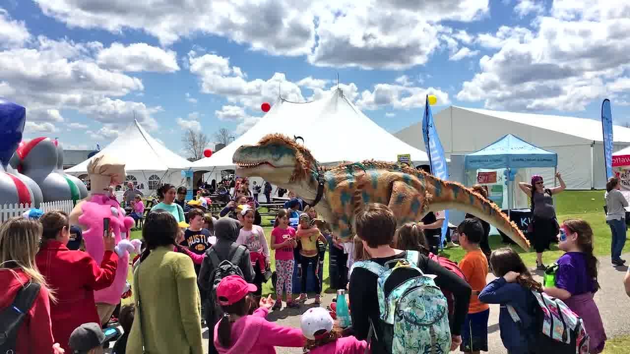 A group of children crowd around a fake dinosaur on a sunny day.