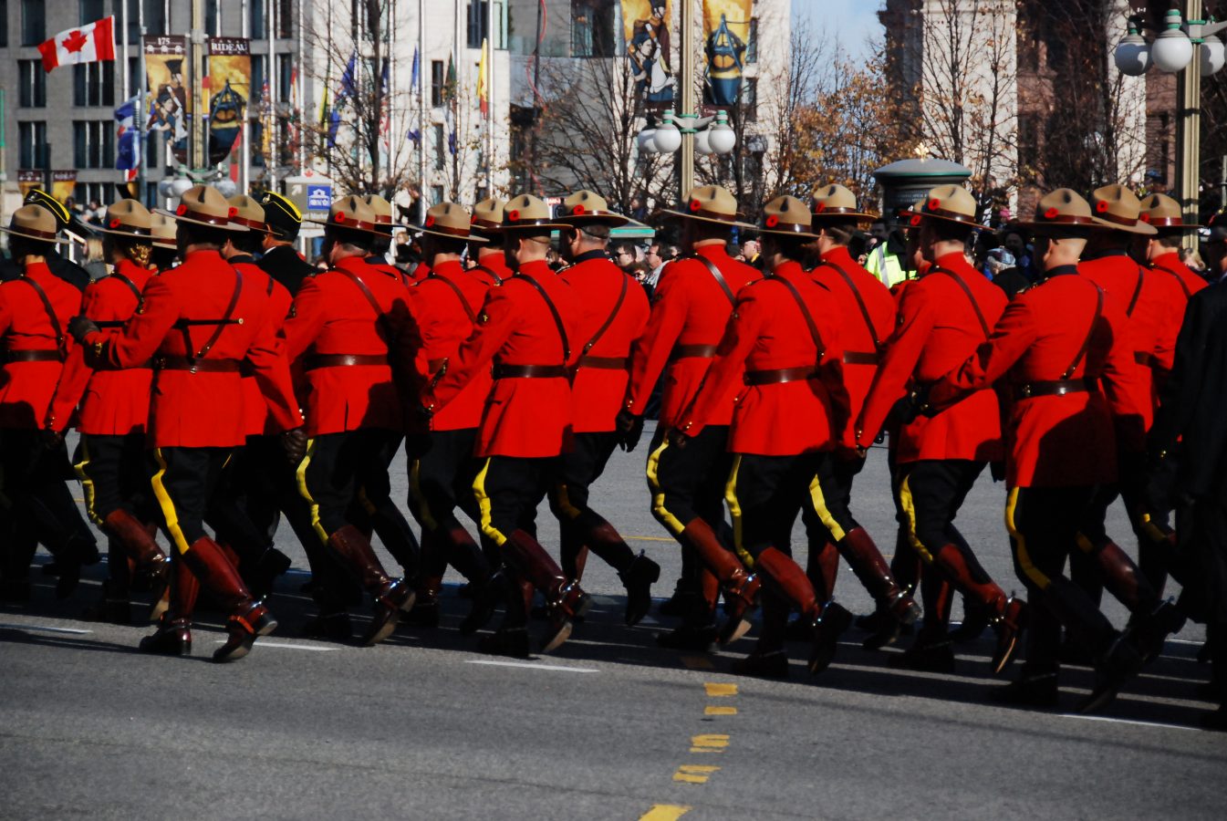The RCMP walk down Elgin Street in Ottawa, dressed in their red coats, blue pants with a yellow stripe on the side, and brown leather knee-high boots.