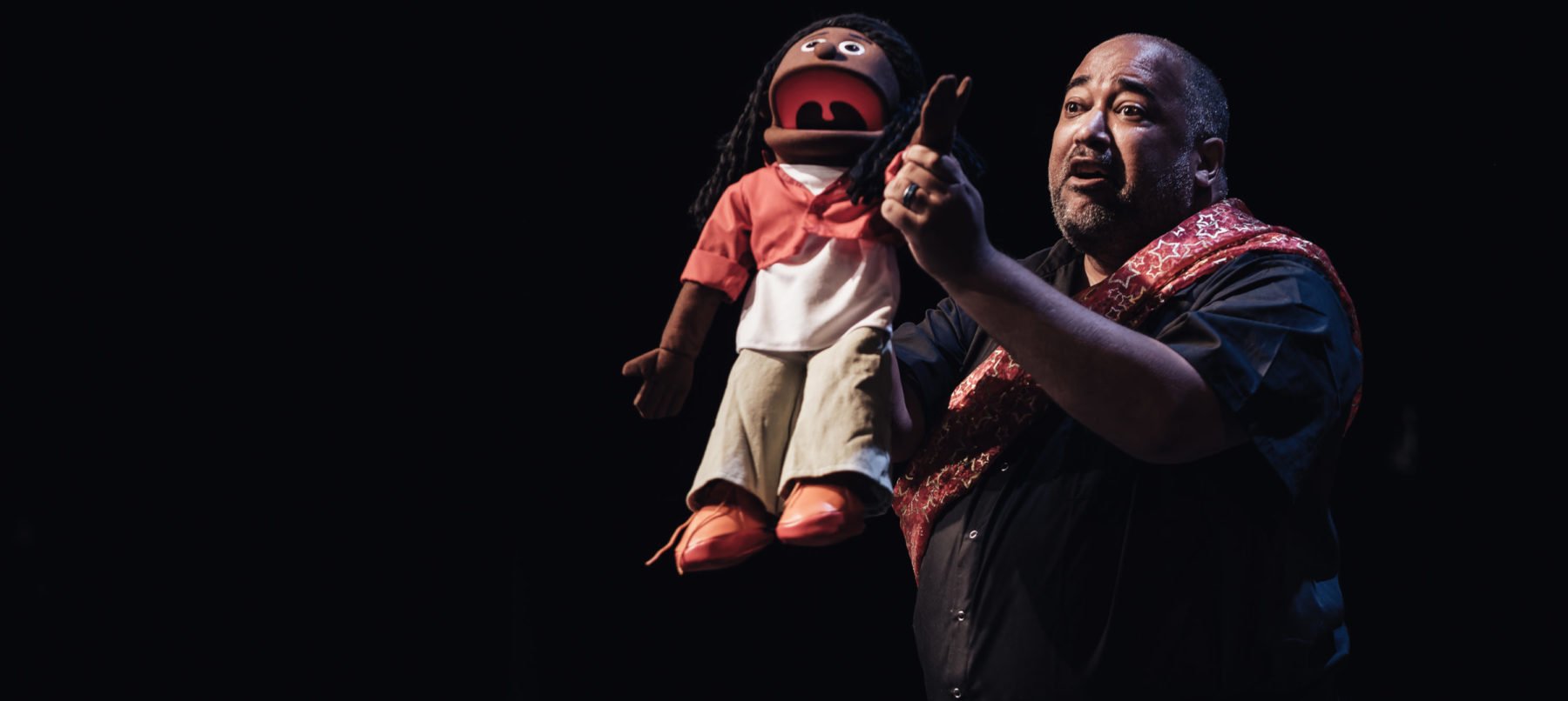 A man holding a puppet on-stage. The puppet's mouth is open as if speaking and his hand is raised.