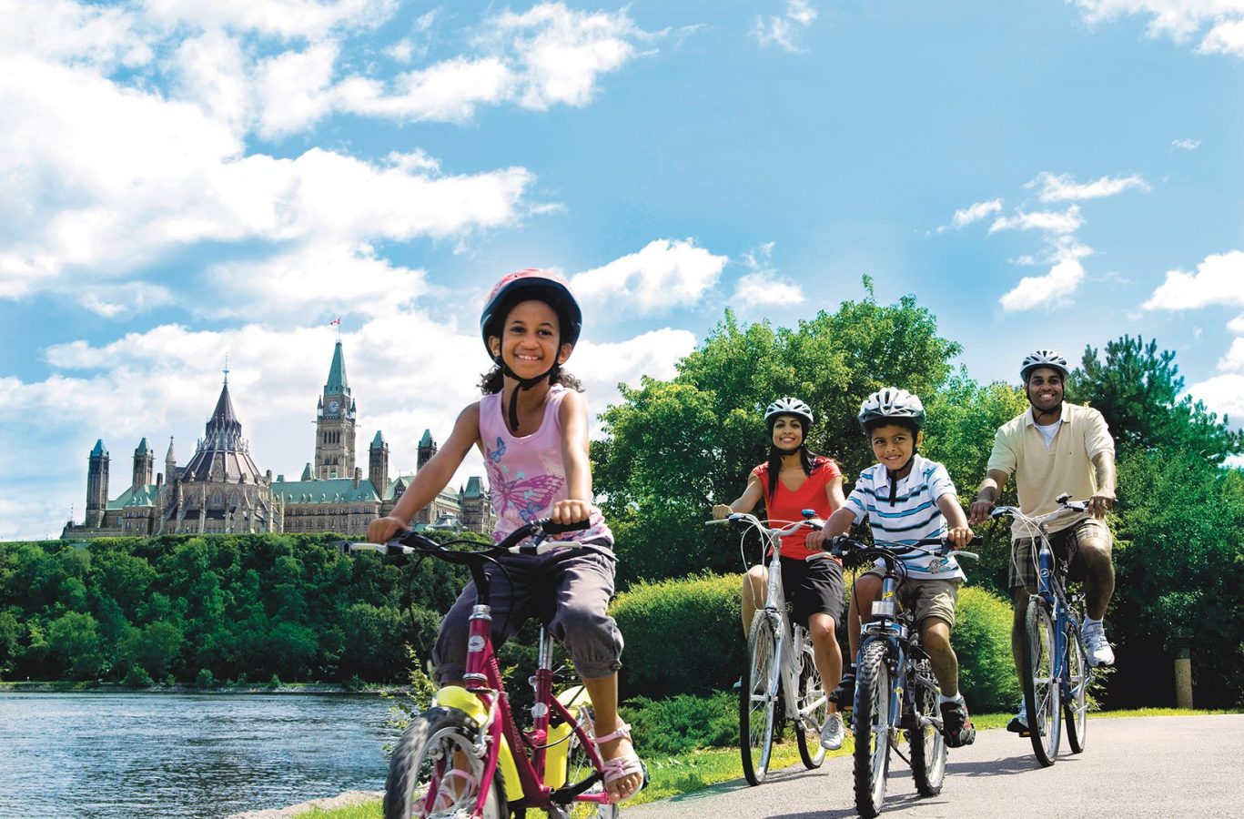 A family biking along the Rideau Canal in Ottawa during the summertime, Parliament Hill in the background.