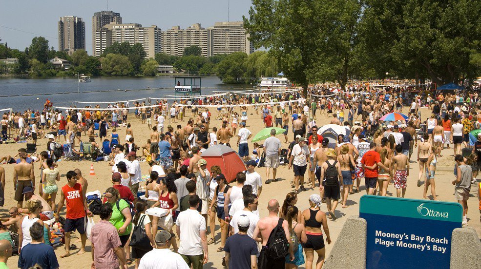 A crowd of people on Mooney's Bay Beach in Ottawa, with beach volleyball courts, colourful umbrellas, and a lifeguard stand.