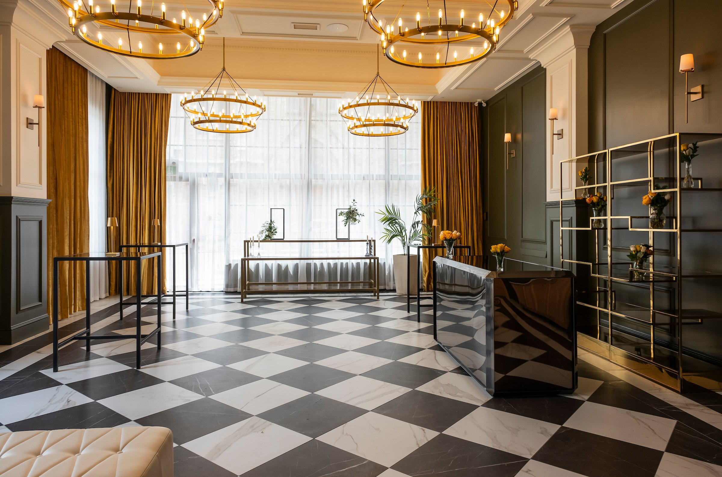 A room with black and white checkered marble floor, floor to ceiling windows, and large gold chandeliers.