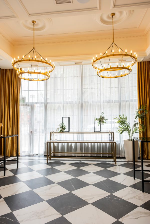 A room with black and white checkered marble floor, floor to ceiling windows, and large gold chandeliers.