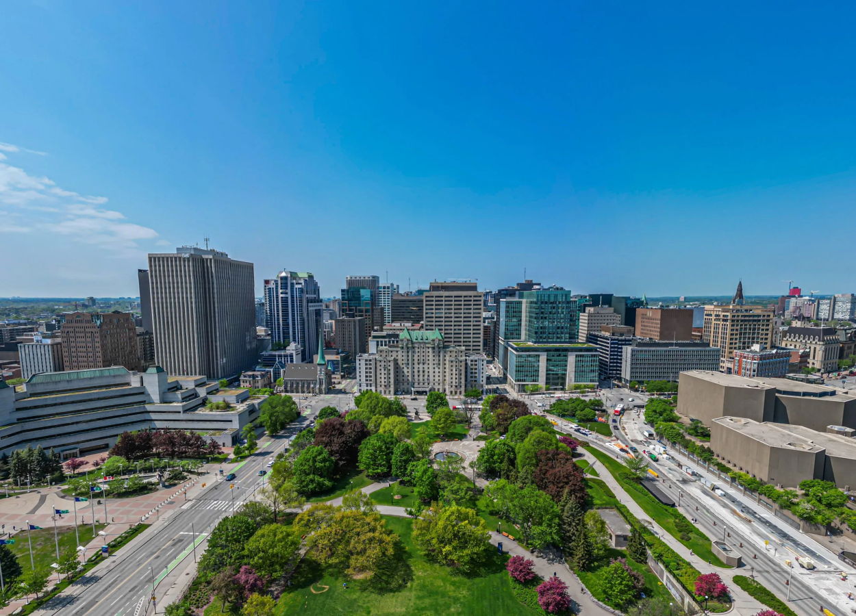 An aerial view of the Lord Elgin Hotel from Confederation Park in the daytime. There are no clouds in the sky, and the grass of the park is bright green.